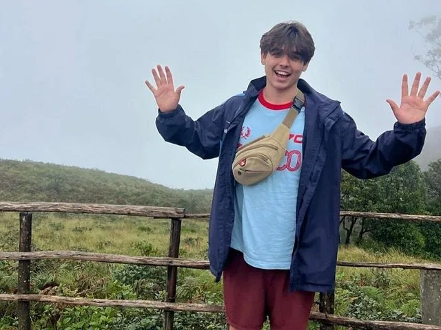 A young man smiling with raised hands outdoors in a misty green landscape.